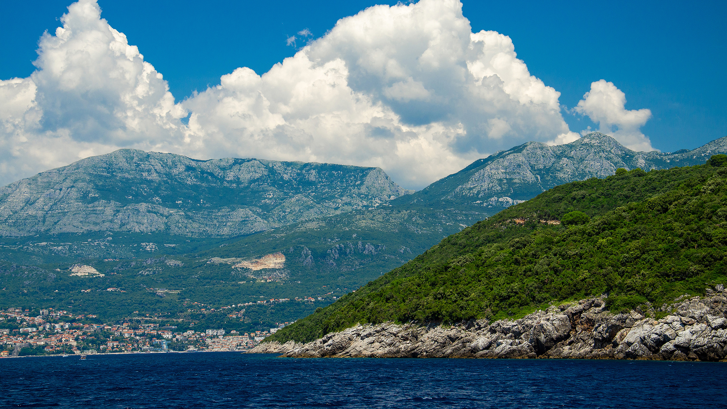 View of Boka Kotor bay water, town of Herceg Novi and Mount Orjen of Dinaric Alps mountain range in front of blue sky with white clouds, Montenegro